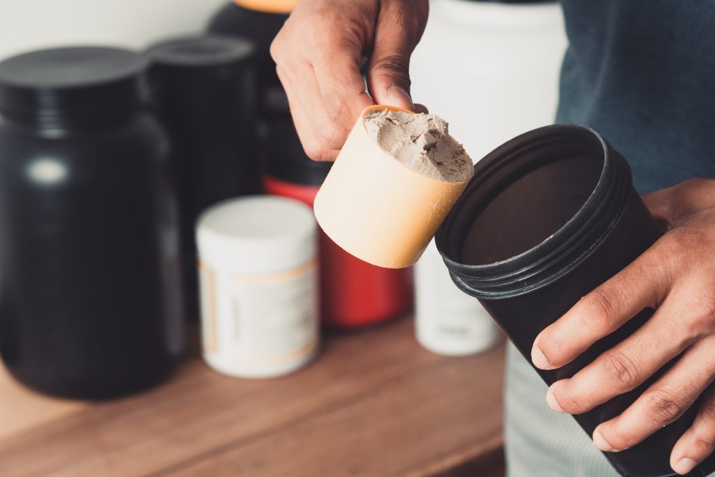 Hands holding scoop of protein powder and shaker bottle.